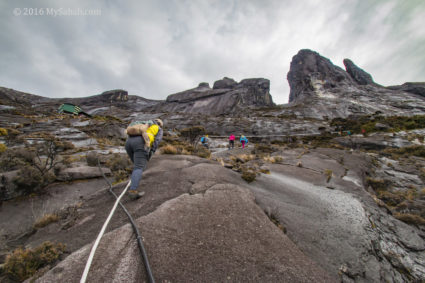 Reaching the Sayat-Sayat Checkpoint (green building at the left). At the right are climbers on Ranau Trail.